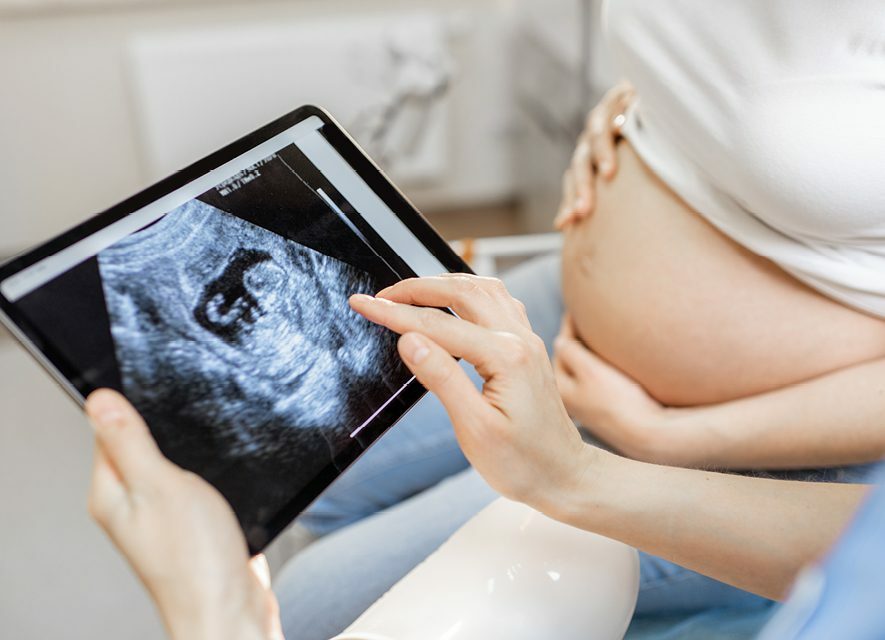 Healthcare professional with digital tablet showing ultrasound image to pregnant woman in the background with her hands on her bare abdomen