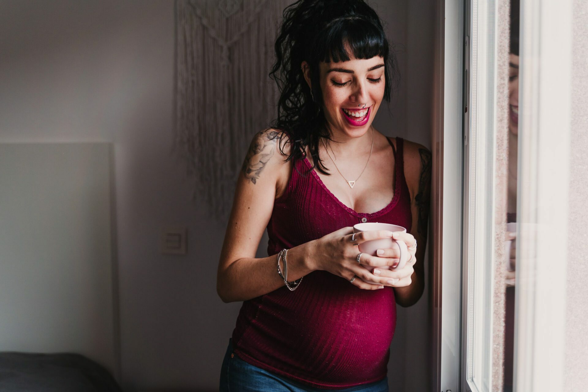 Pregnant woman smiling as she holds a mug and stands by the window