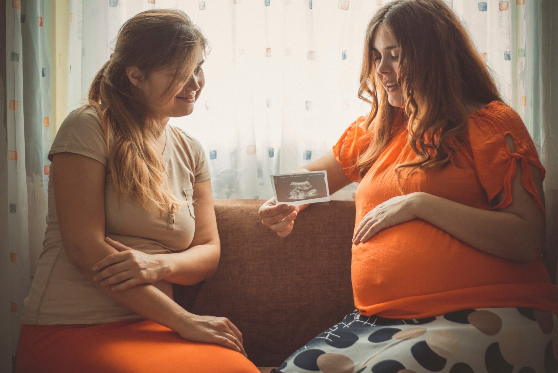 Pregnant woman sitting with female friend and showing photo of ultrasound